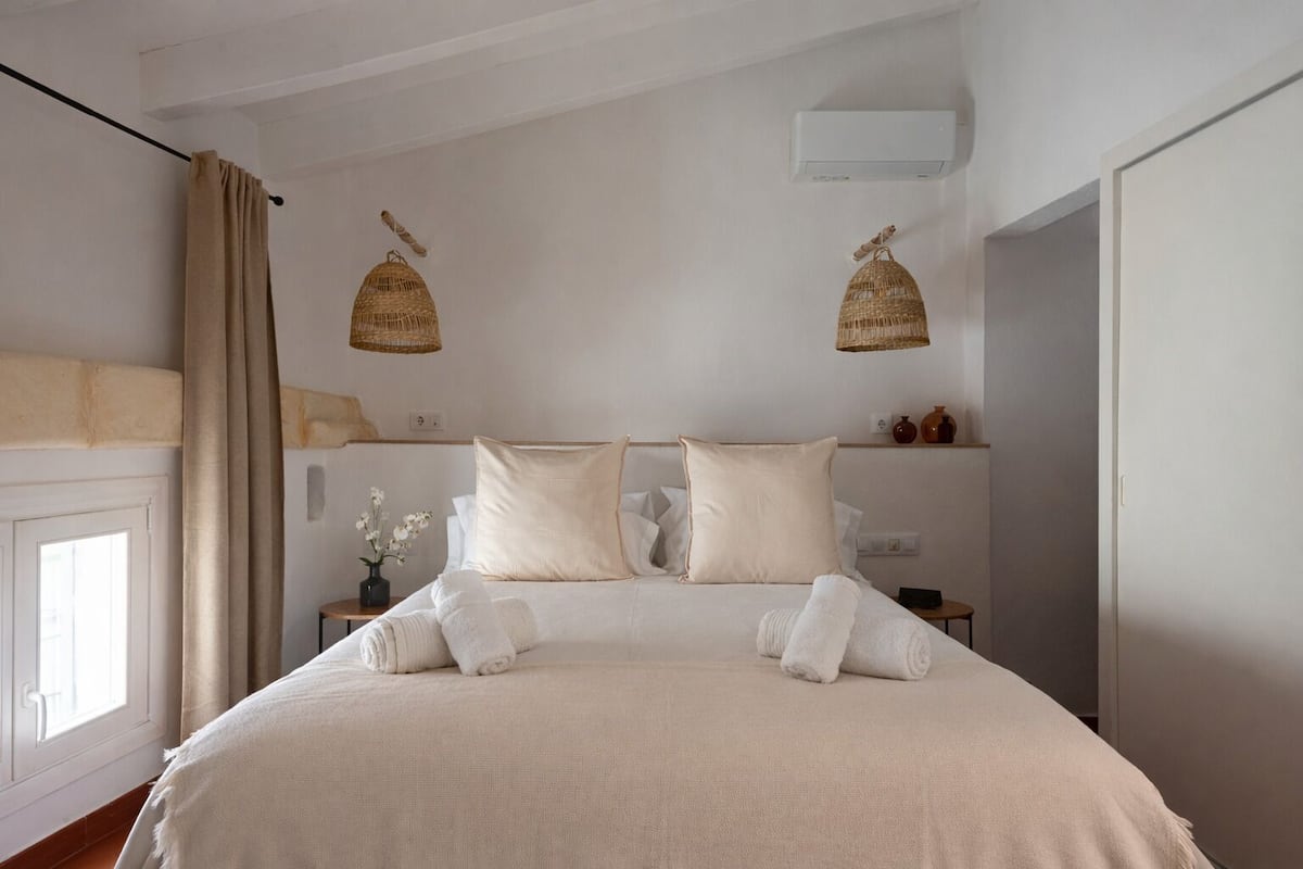 Une chambre à coucher avec un grand lit, deux tables de chevet, une petite table, une lampe en osier et un climatiseur fixé au mur.