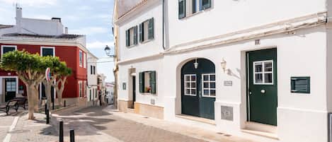 A narrow street lined with traditional buildings, featuring a mix of white and red facades, a cobblestone path, and a tree with green foliage.