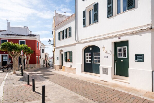 Une rue étroite bordée de bâtiments traditionnels, avec un mélange de façades blanches et rouges, un chemin pavé et un arbre aux feuilles vertes.