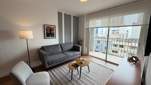 A modern living room with a grey sofa, a small round table, and a floor lamp. A framed picture hangs on the wall, and a large window offers a view of the cityscape. - w Chic Inviting 2BR w Balcony in Miraflores (Lima)