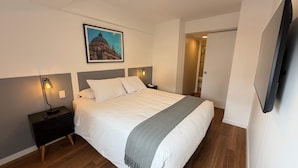 A neatly made bed with white linens and a grey throw, flanked by bedside tables with lamps, a framed picture above the bed, and a TV mounted on the wall. - w Chic Inviting 2BR w Balcony in Miraflores (Lima)