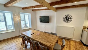 A dining area with a wooden table and chairs, a TV mounted on the wall, and a large window with blinds. - Lush Sanctuary Near Forest (Mont-Dore)