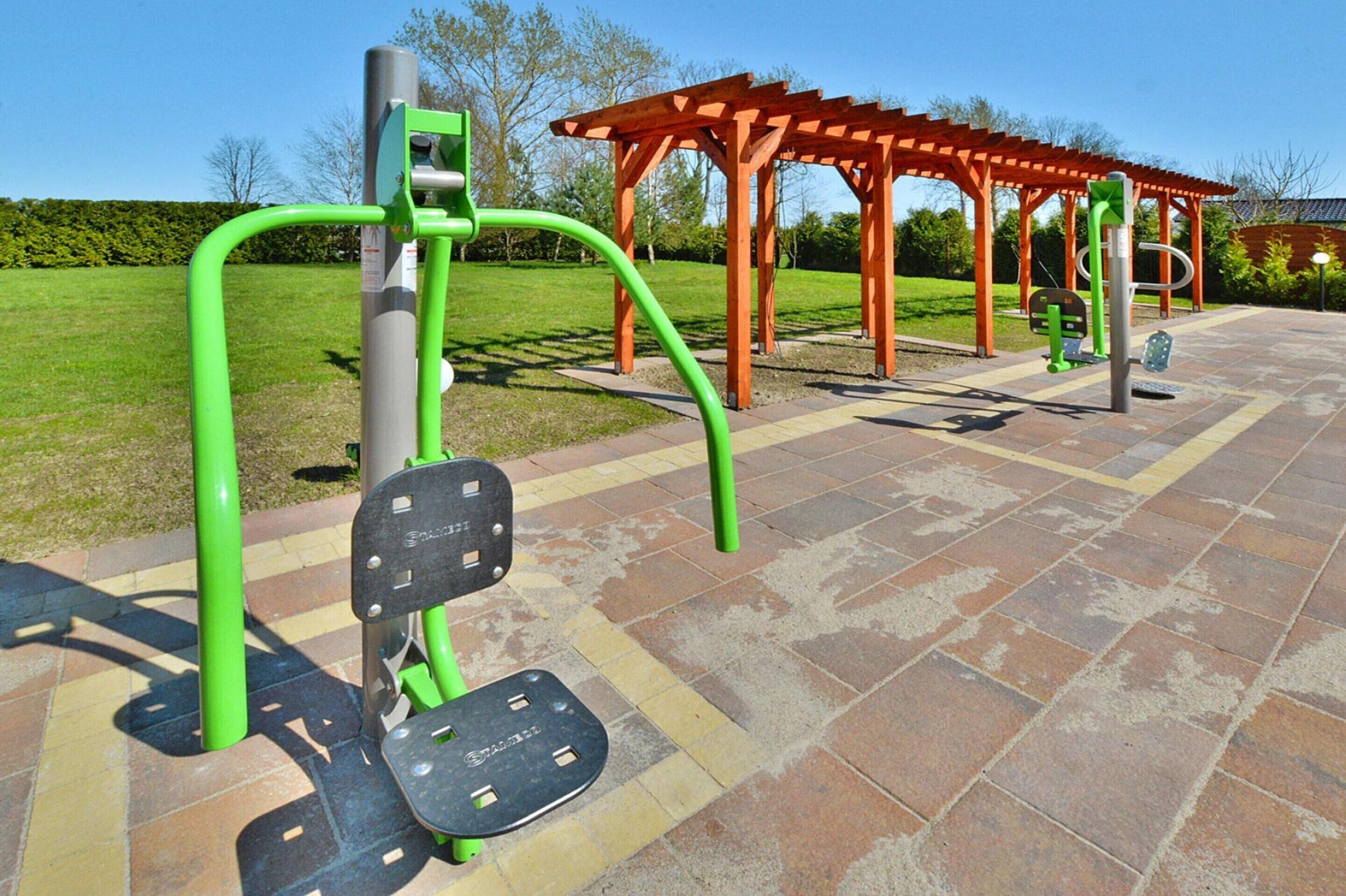 Outdoor exercise equipment, a wooden pergola, and a paved area with a hedge in the background.