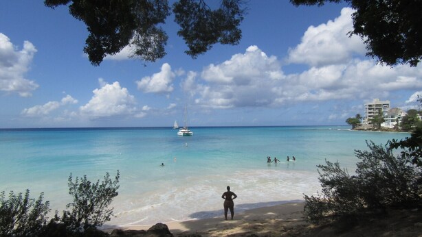 Una playa con agua turquesa cristalina, una persona de pie en la orilla y un barco a lo lejos.