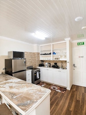 A compact kitchen with white cabinets, a stainless steel refrigerator, a microwave, and a stove. There is a wooden floor and a patterned rug. - 202 Batts Rock Beach Condos 1 Bedroom Suite (Bridgetown)