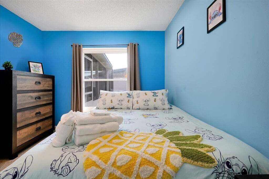 Een slaapkamer met een blauwe muur, een raam met gordijnen, een bed met beddengoed met tekenfilms, een houten dressoir en een aan de muur hangend schilderij.