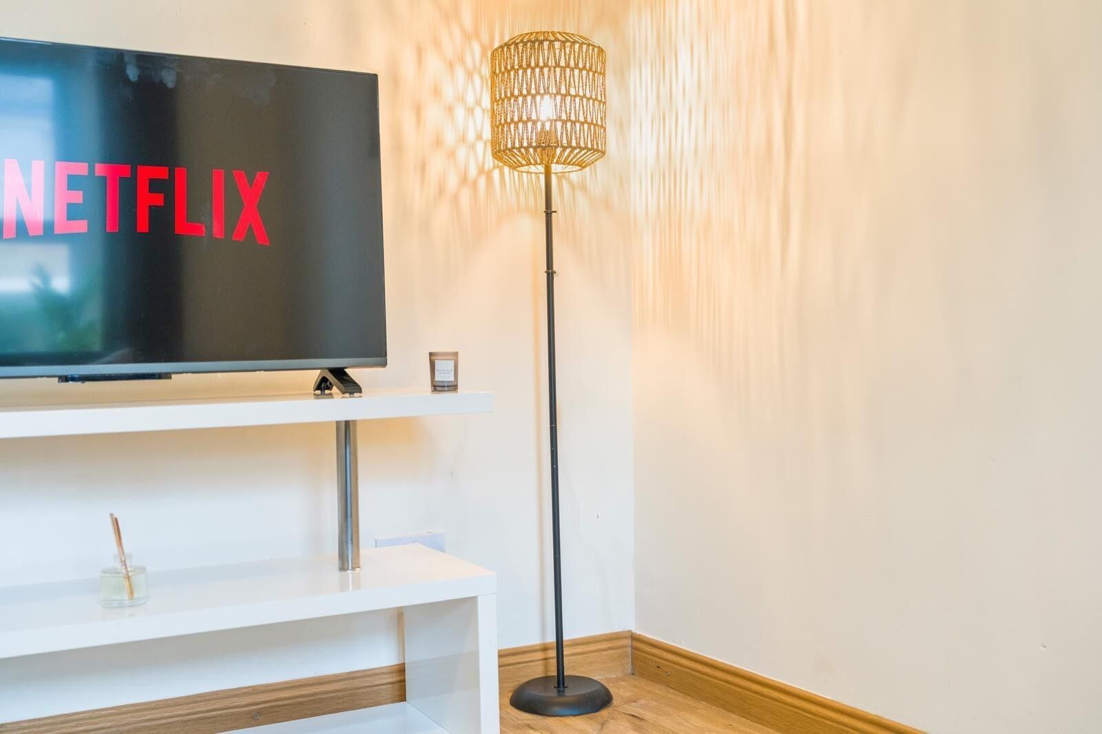 A modern living room with a flat-screen TV on a white stand, a floor lamp, and a small table with a candle.