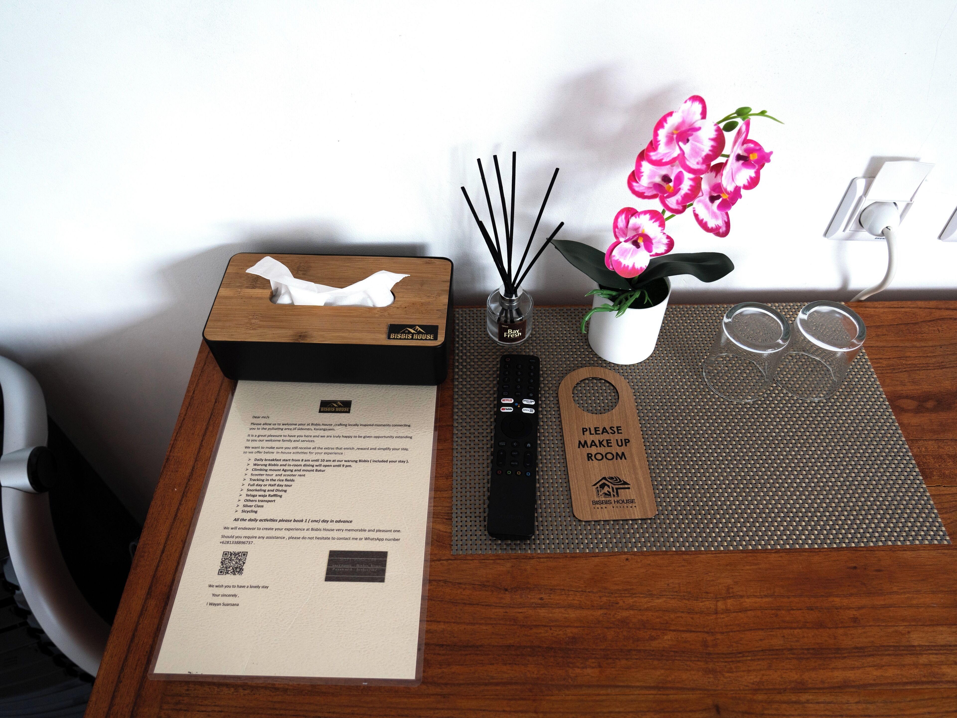 A wooden table with a tissue box, a remote control, a flower vase, a pair of glasses, and a placard reading "PLEASE MAKE UP ROOM".