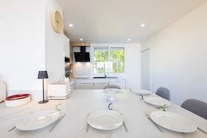 A modern dining area with a white table set for four, a kitchen in the background, and a window with a view of trees.