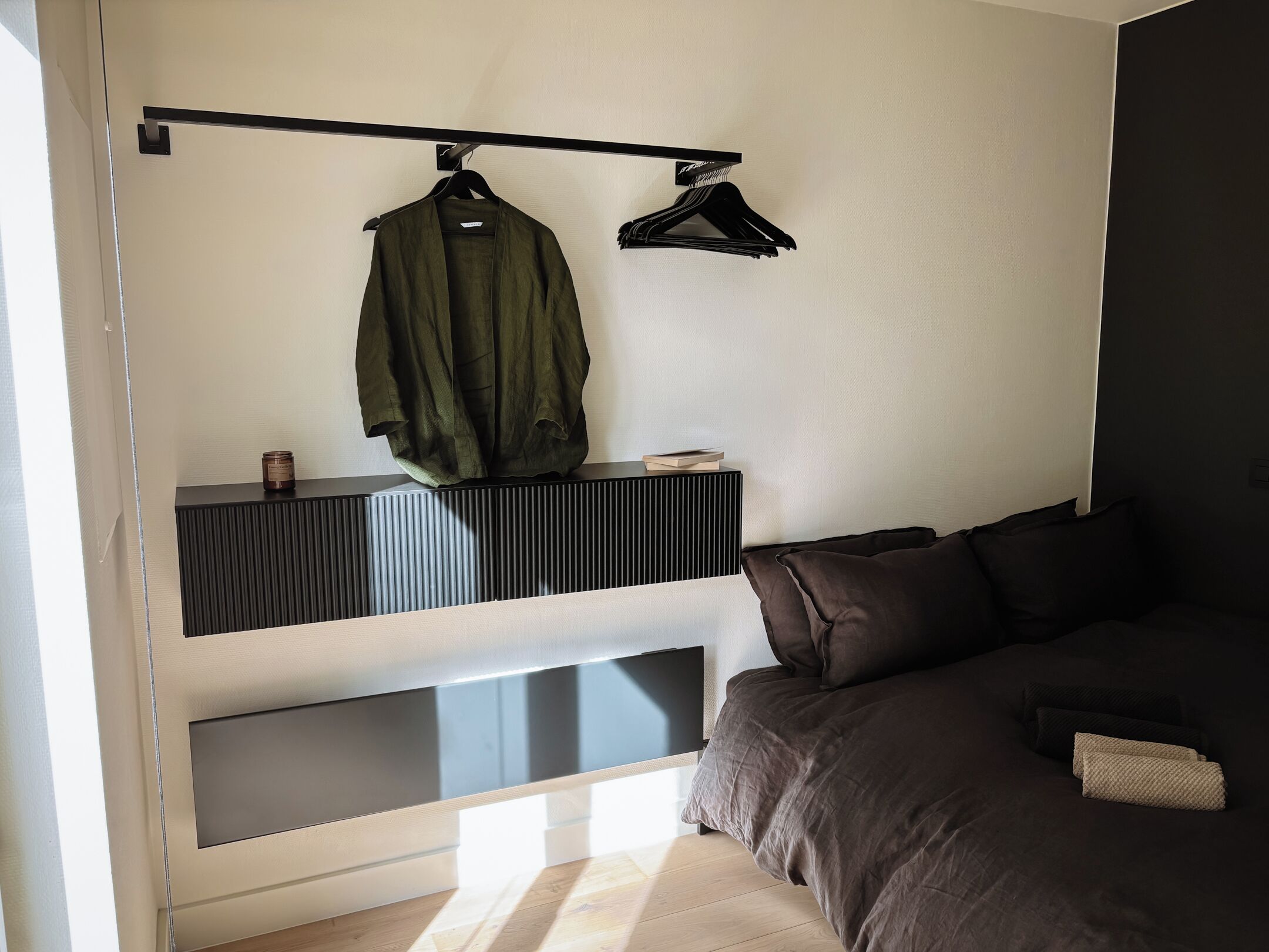 Une chambre moderne avec un lit, un porte-manteau suspendu et une petite étagère sur laquelle se trouvent un livre et une bougie.