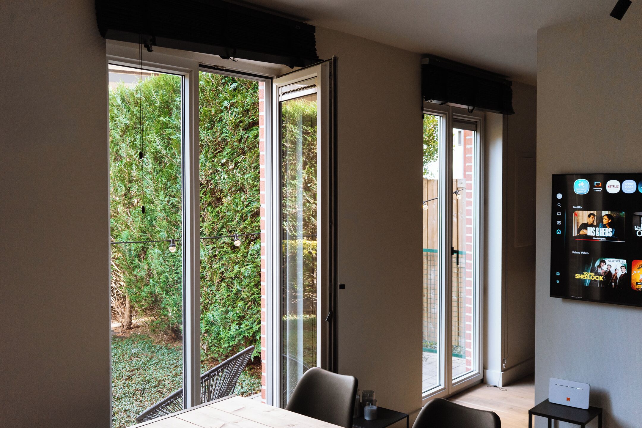 A room with a sliding glass door to a garden, a flat-screen TV mounted on the wall, and a small table with a laptop.