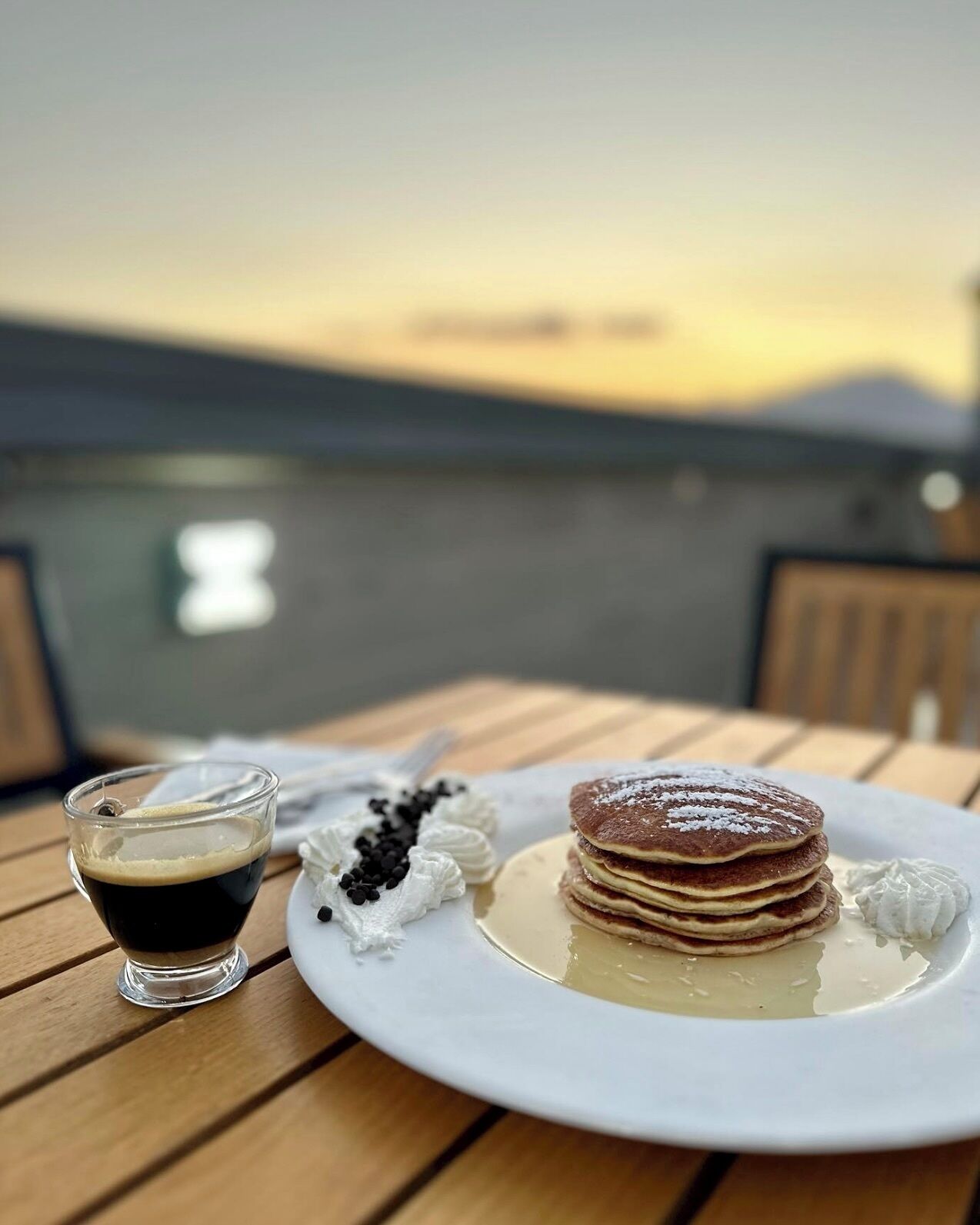 A breakfast setting with pancakes, whipped cream, and a cup of coffee on a wooden table with a scenic view in the background.