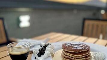 A breakfast setting with pancakes, whipped cream, and a cup of coffee on a wooden table with a scenic view in the background.