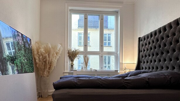Een moderne slaapkamer met een groot bed, een decoratieve plafondlamp, een raam met uitzicht op gebouwen en een visgraat parketvloer.