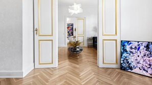 Een hal met houten vloer, witte muren en deuren met gouden lijst. Een groot televisiescherm is aan de muur bevestigd.