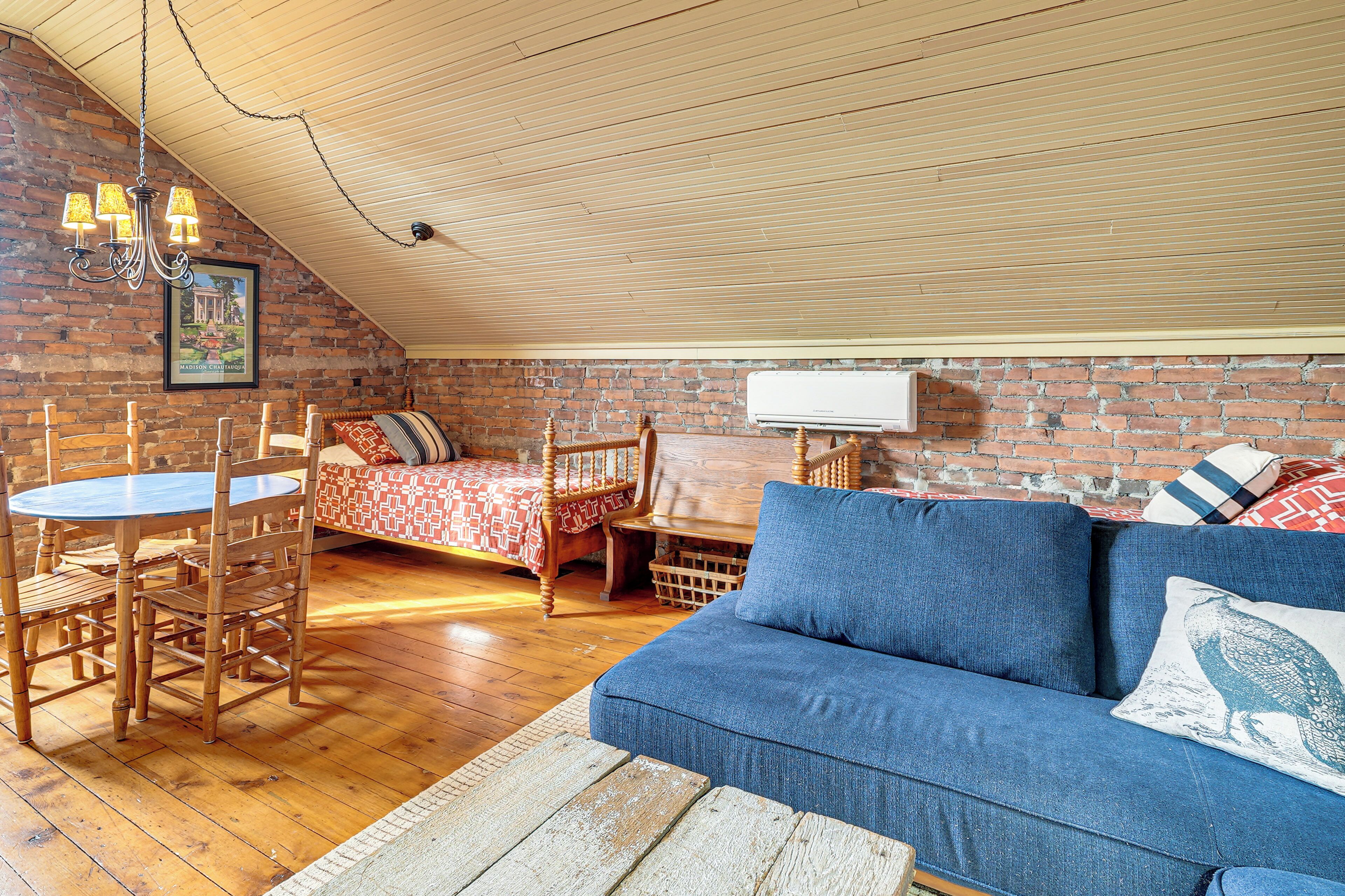 A loft-style room with a wooden ceiling, a dining table with chairs, a sofa, and a bed with patterned bedding.