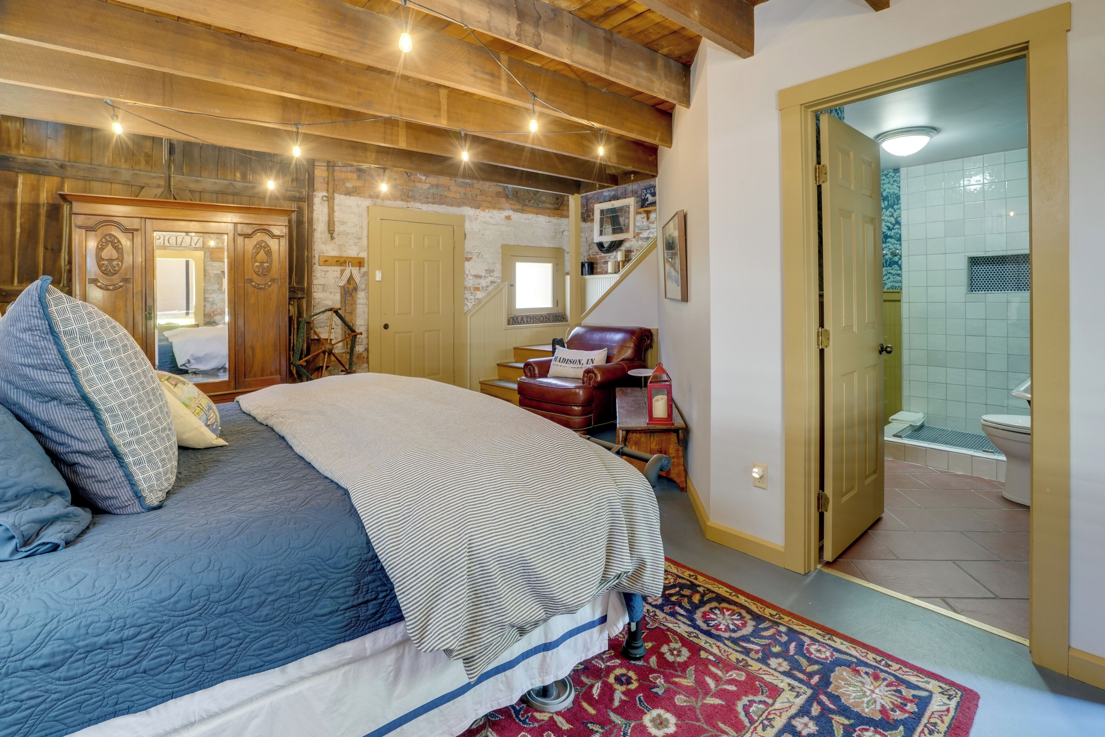 A bedroom with a bed, a wooden wardrobe, a small table with a lamp, and a bathroom visible through an open door.