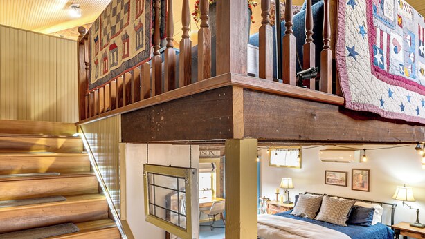A wooden staircase with a view of a bedroom below, featuring a bed, nightstands, and a quilt.
