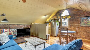 A cozy living room with a blue sofa, a wooden coffee table, and a fireplace. There is a kitchen area with a refrigerator and a dining table with chairs. The room has a wooden ceiling and brick walls.