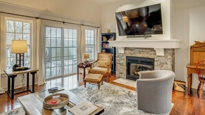 A spacious living room with a fireplace, a television mounted above it, and a large window with curtains. - Canoe, Swim & Hike: Lakefront Brandenburg Home! (Brandenburg)