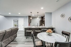 A modern kitchen with dark cabinets, a central island, pendant lighting, and a dining area with a white marble table and leather chairs. - Easy Commute to Philly: Traveling Pros' Hub! (Levittown)
