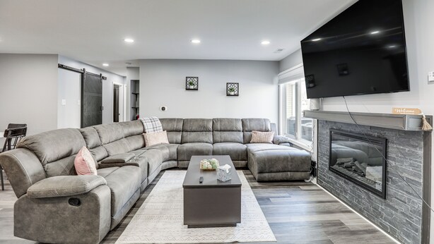 A modern living room with a sectional sofa, a flat-screen TV mounted above a fireplace, and a small table with decorative items. - Easy Commute to Philly: Traveling Pros' Hub! (Levittown)