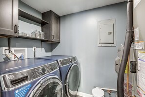 A laundry room with a blue front-loading washer and dryer, a shelf with storage containers, and a utility meter on the wall. - Easy Commute to Philly: Traveling Pros' Hub! (Levittown)