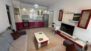 En moderne stue med sofa, sofabord, fjernsyn og et køkkenområde med skabe og et køleskab.