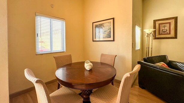 A dining area with a round table, four chairs, two windows with blinds, and a framed painting on the wall. - Serena Stay Spacious 3BR Apartment (Puerto Peñasco)