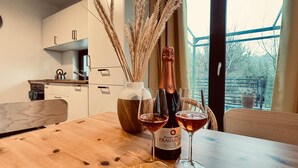 Una mesa de madera con dos copas de vino y una botella de vino Fragolino Duchessali. Hay un florero con plantas secas y una ventana con vistas a árboles.