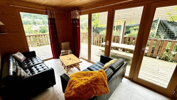A cozy room with a sofa, a table, and a view of a wooden deck and greenery outside. - Spacious French Chalet Stay for 10 (Mont-Dore)