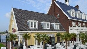 A waterfront hotel with a yellow facade, white-framed windows, and a terrace with outdoor seating. - Explore Huizen in Perfect Suite w/Onsite Dining Near Breathtaking Reserves (Huizen)