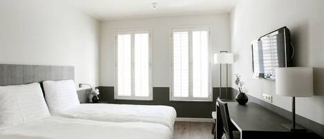 A hotel room with a large bed, a desk, a chair, a lamp, and two windows.