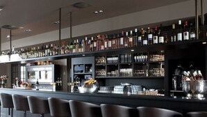 Zona de bar con varios taburetes, una barra con botellas y vasos, y un refrigerador.