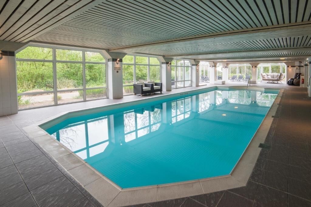 An indoor swimming pool with clear windows, lounge chairs, and a tiled floor.