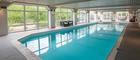 An indoor swimming pool with clear windows, lounge chairs, and a tiled floor.