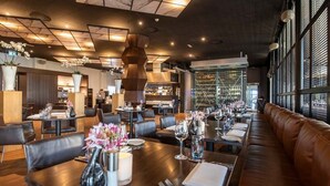 Interior de restaurante moderno com mesas de madeira escura, assentos de couro e uma adega ao fundo.