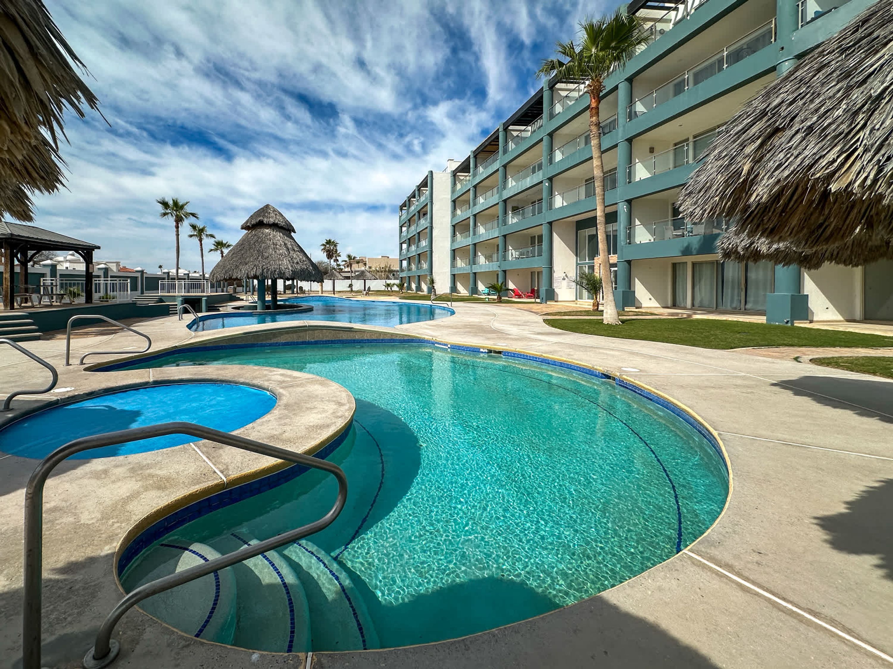 Zona de piscina en un resort con varias piscinas, palmeras y un edificio con balcones.