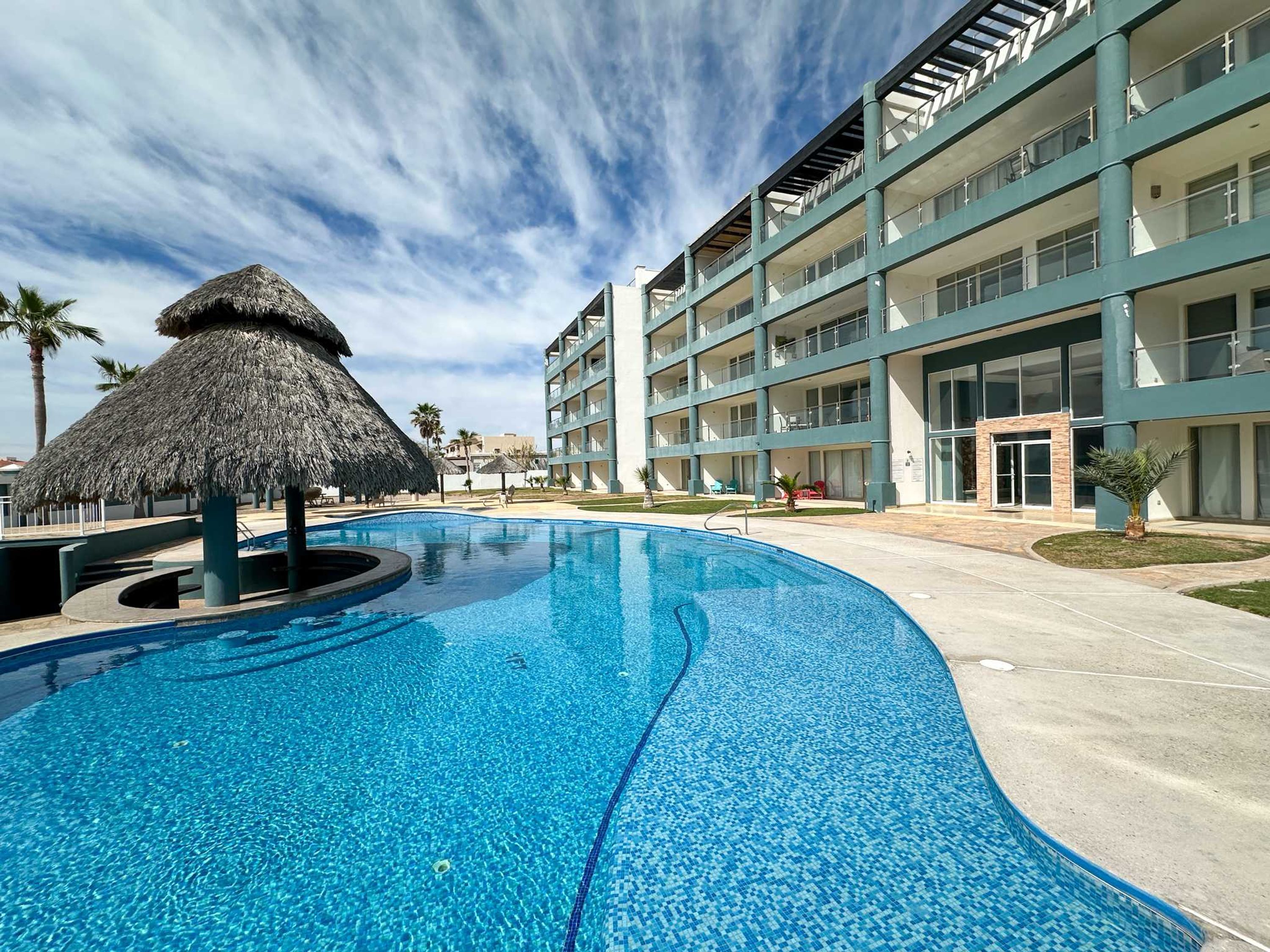Una piscina con una estructura de techo de paja, un edificio de varios pisos con balcones y un cielo despejado.