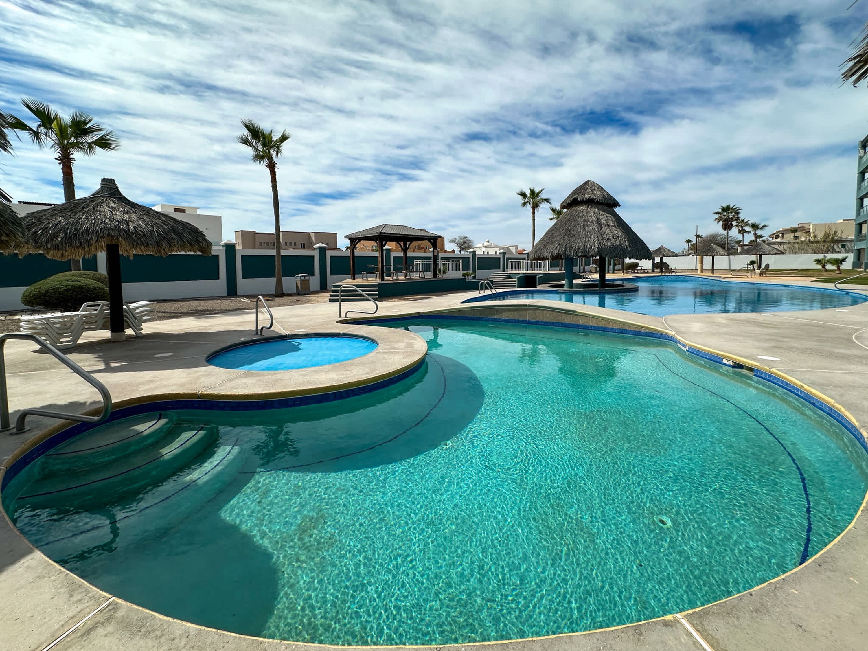 Una piscina cristalina con una zona hundida en el centro, rodeada de sillas de descanso y sombrillas de paja.