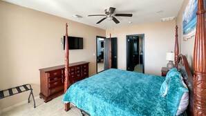 Een slaapkamer met een hemelbed, een plafondventilator, een dressoir, een televisie en een deur die naar een andere kamer leidt.
