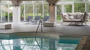 Un espace piscine intérieure doté de chaises longues et d’un canapé, le tout entouré de grandes fenêtres.