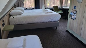 Une chambre d’hôtel avec un lit, un bureau et une chaise. On y trouve une fenêtre avec des rideaux, un téléphone fixé au mur et un tableau d’affichage.