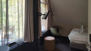 A hotel room with a bed, a chair, a small table, a round ottoman, and a window with curtains.