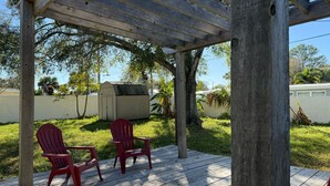 En överbyggd träveranda med två röda Adirondack-stolar, ett litet förråd och en vit byggnad i bakgrunden.