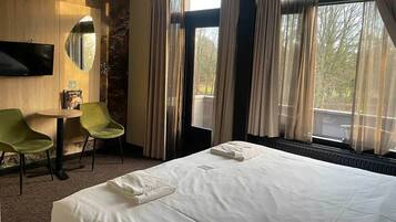 A hotel room with a bed, two green chairs, a small table, a TV, and a window with curtains.