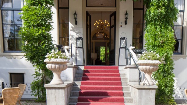 Une entrée imposante, avec un tapis rouge menant à un escalier, de part et d’autre duquel se trouvent de grands jardinières et un lustre au-dessus de la porte.