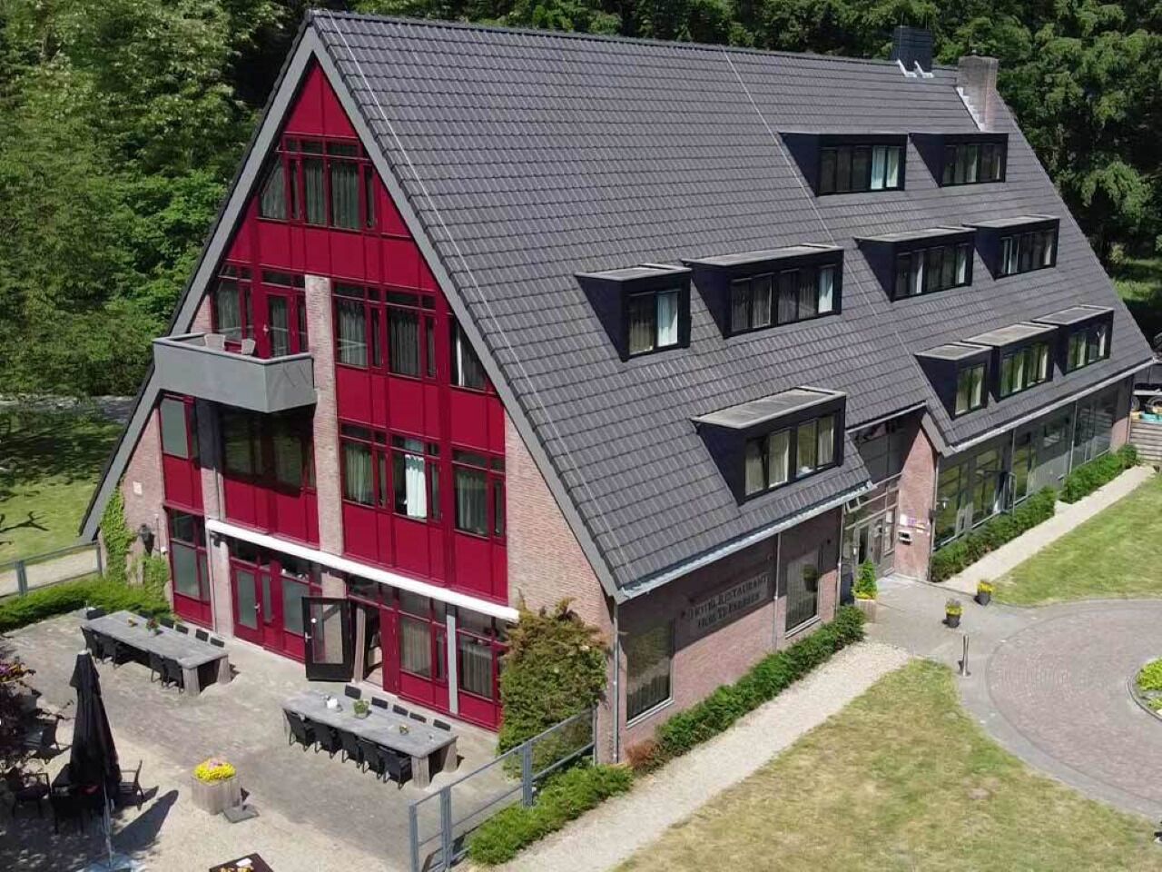 A two-story building with a red facade and grey roof, featuring multiple windows and a paved outdoor area with tables and chairs.