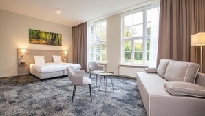 Une chambre d’hôtel comprenant un lit, deux fauteuils, un canapé, une petite table et une grande fenêtre avec des rideaux.