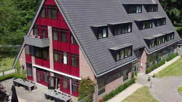 A two-story building with a red facade and grey roof, featuring multiple windows and a paved outdoor area with tables and chairs.
